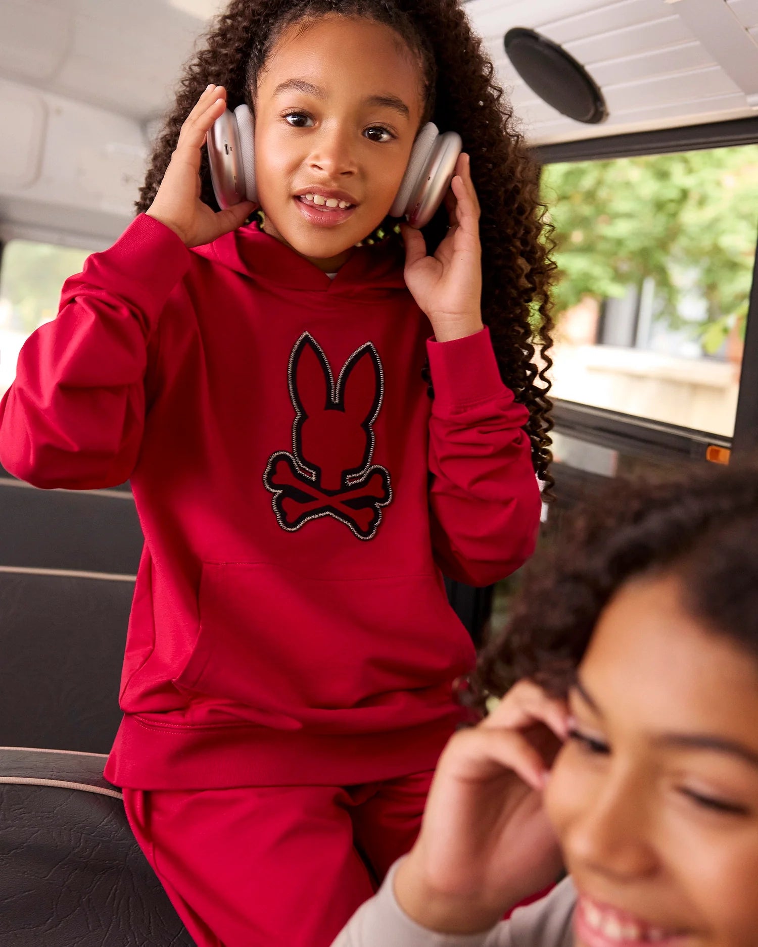 A young girl with long curly hair, wearing a red hoodie with a bunny-and-crossbones logo and KIDS WALTER LIGHTWEIGHT SWEATPANT - B0P860D200 by Psycho Bunny, sits on a bus seat while adjusting her large headphones. Another young girl in the foreground, slightly out of focus, is smiling.