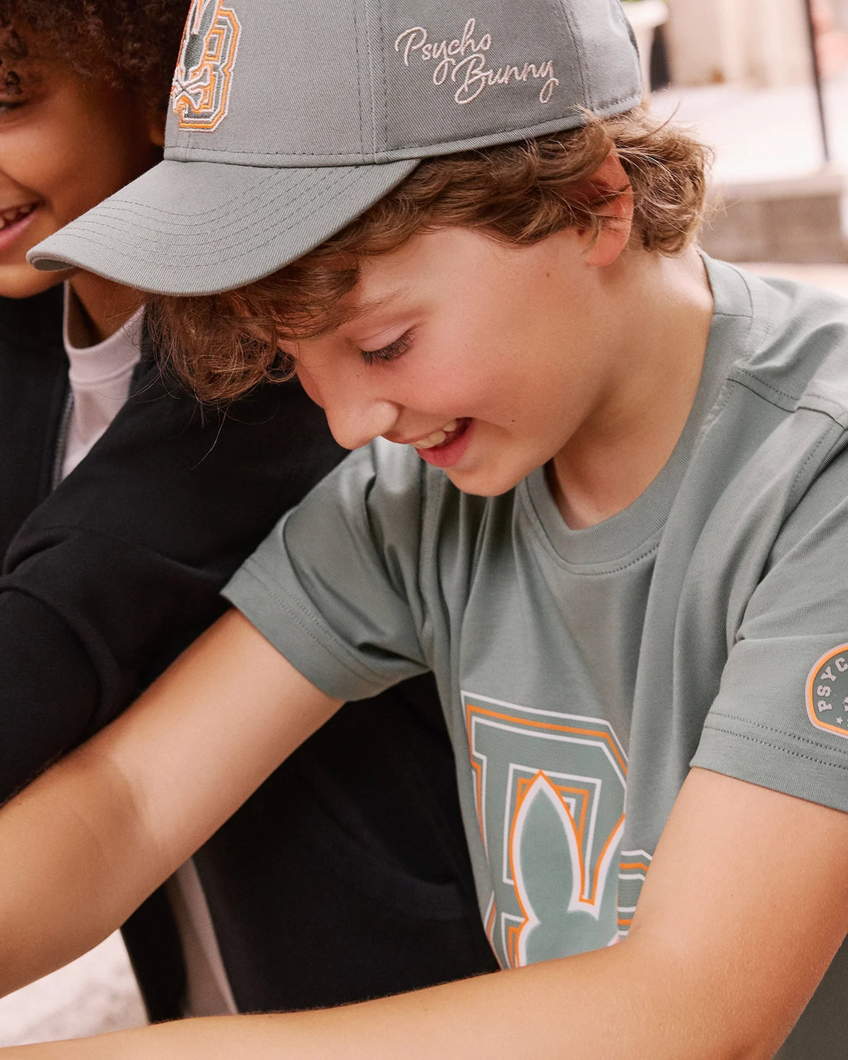 A young boy is smiling as he wears the KIDS ZACK BASEBALL CAP from Psycho Bunny in gray, along with a T-shirt featuring their stylized bunny logo. He's outdoors on a sunny day, leaning over something out of frame. The woven cotton of his cap enhances his sporty look, while another person is partially visible beside him.