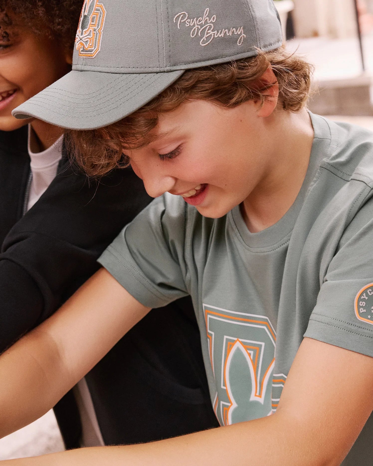 A young boy is smiling as he wears the KIDS ZACK BASEBALL CAP from Psycho Bunny in gray, along with a T-shirt featuring their stylized bunny logo. He's outdoors on a sunny day, leaning over something out of frame. The woven cotton of his cap enhances his sporty look, while another person is partially visible beside him.
