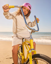 A young child with long curly hair and a pink cap rides a yellow bike on a beach. Dressed in Psycho Bunny's KIDS LYONS SWEATSHORT - B0R578C200 and a beige sweatshirt, the child pedals happily as the ocean waves crash under the blue sky in the background.