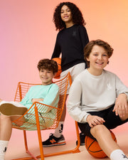 Three children pose with basketballs against a gradient background. One sits on an orange chair, sporting a mint green outfit made from premium cotton fabric. The others stand, dressed in the KIDS HOUSTON FRENCH TERRY CREWNECK by Psycho Bunny in black and light gray, all smiling in their casual sneakers.