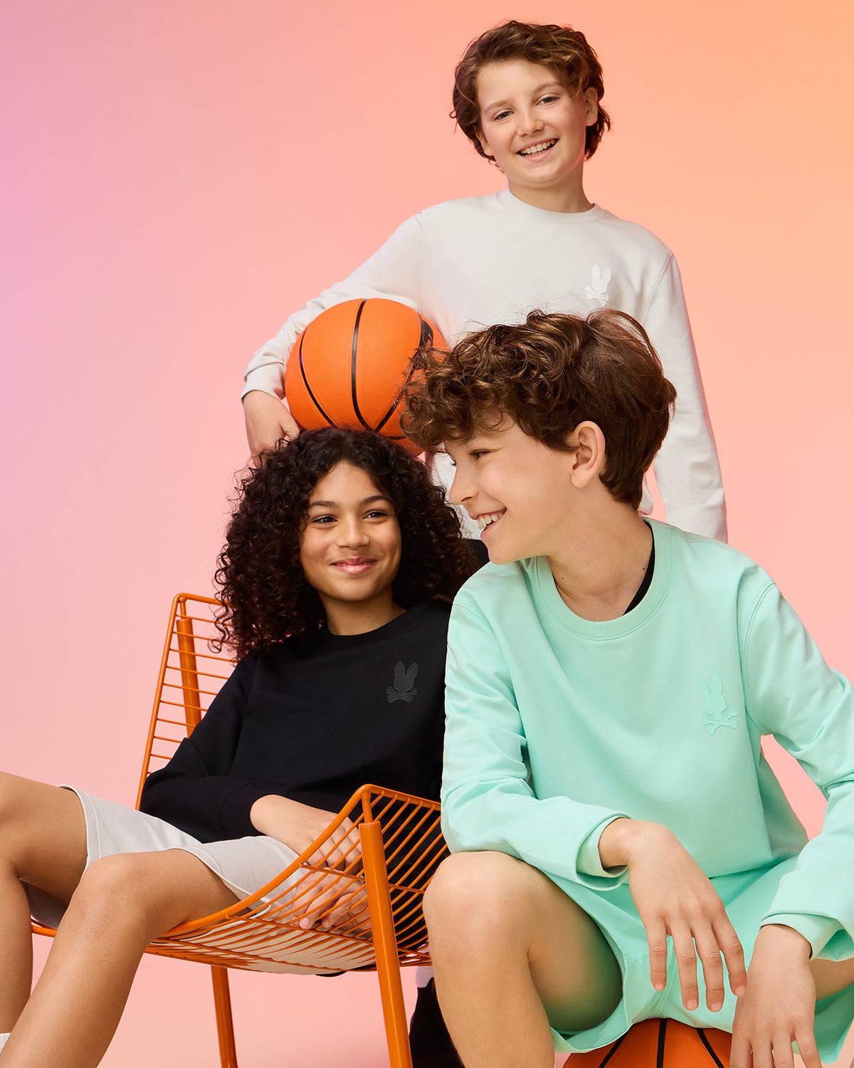 Three kids in casual Psycho Bunny apparel, featuring the KIDS HOUSTON FRENCH TERRY CREWNECK crafted from premium cotton fabric, pose with a basketball. One stands holding the ball while the other two sit and smile against a backdrop of orange and pink gradient hues.