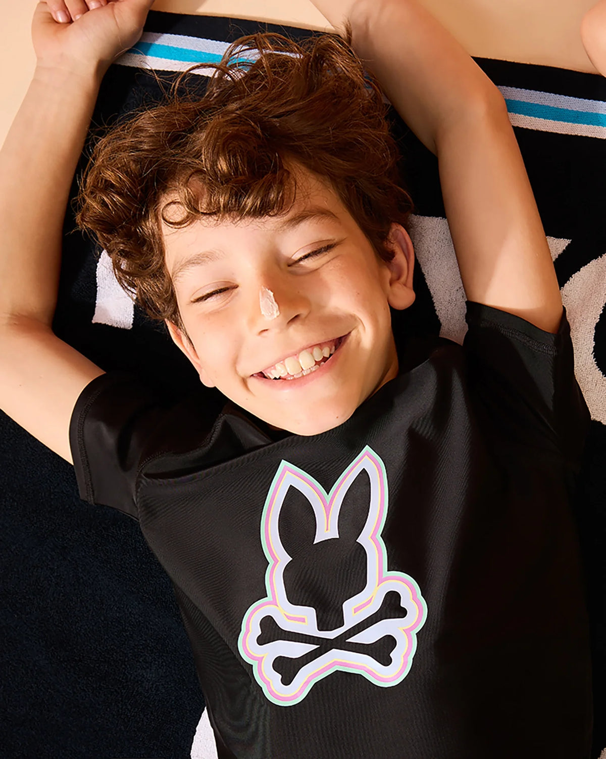 A joyful young boy with curly hair, lying on his back on a striped blanket, smiling with his eyes closed. He wears a Psycho Bunny KIDS SAN LEON ANTI-UV RASHGUARD - B0T645C200 with UPF 50+ protection.