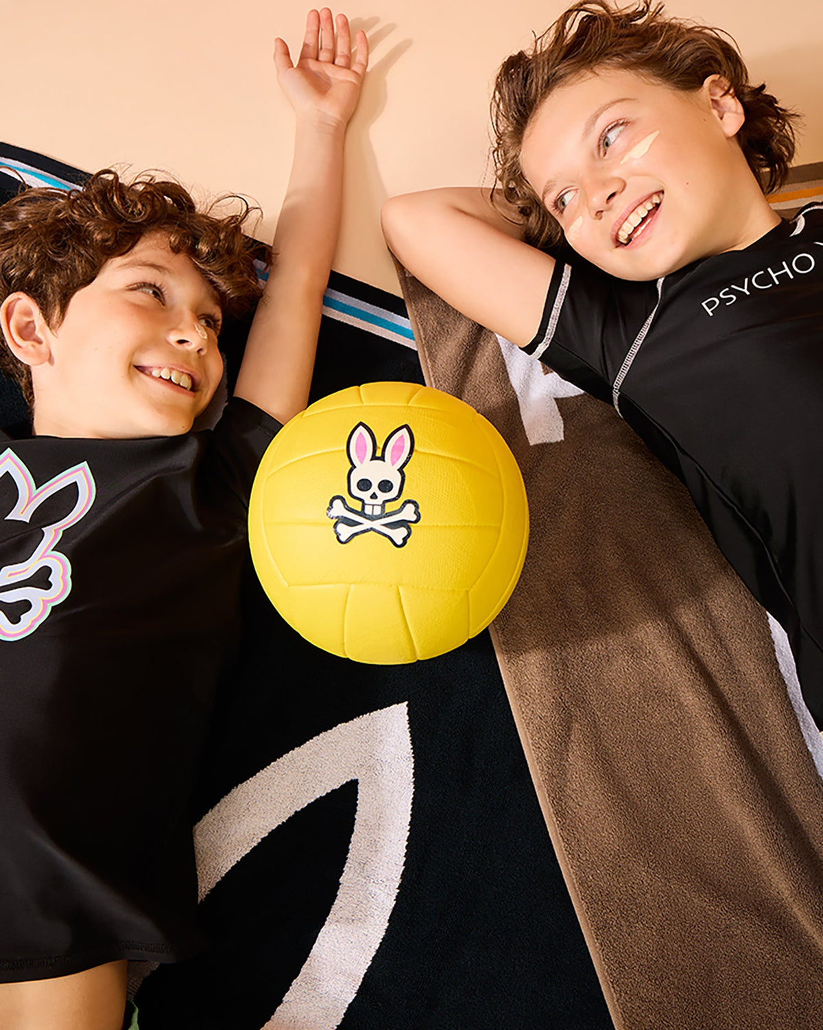 Two children lying on their backs on a colorful blanket, smiling at each other. A bright yellow ball with a pink bunny skull and crossbones design is placed between them. Clad in short sleeve rashguards, specifically the KIDS SHORT SLEEVE ANTI-UV RASHGUARD - B0U558C200 by Psycho Bunny, they appear to be happy and playful under the sun's rays, benefiting from UPF 50+ protection.