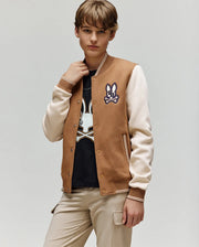 A young person wears the Psycho Bunny KIDS WADE KNIT BOMBER (KO1100001), a varsity-style brown and cream jacket, over a black t-shirt with a bunny graphic and beige pants, standing against a plain light gray background.
