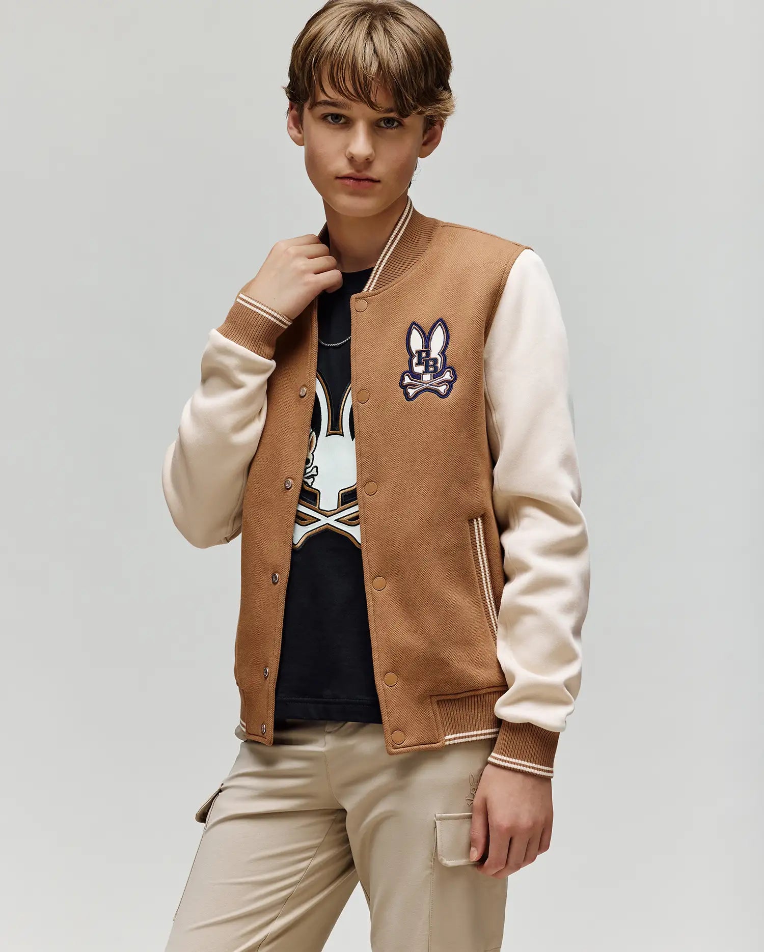 A young person wears the Psycho Bunny KIDS WADE KNIT BOMBER (KO1100001), a varsity-style brown and cream jacket, over a black t-shirt with a bunny graphic and beige pants, standing against a plain light gray background.