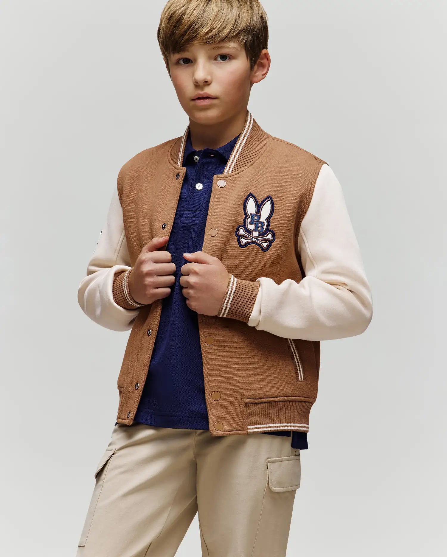 A boy with light brown hair wears the Psycho Bunny KIDS WADE KNIT BOMBER (KO1100001) in tan and white, paired with a navy shirt and beige pants, standing against a plain light background.