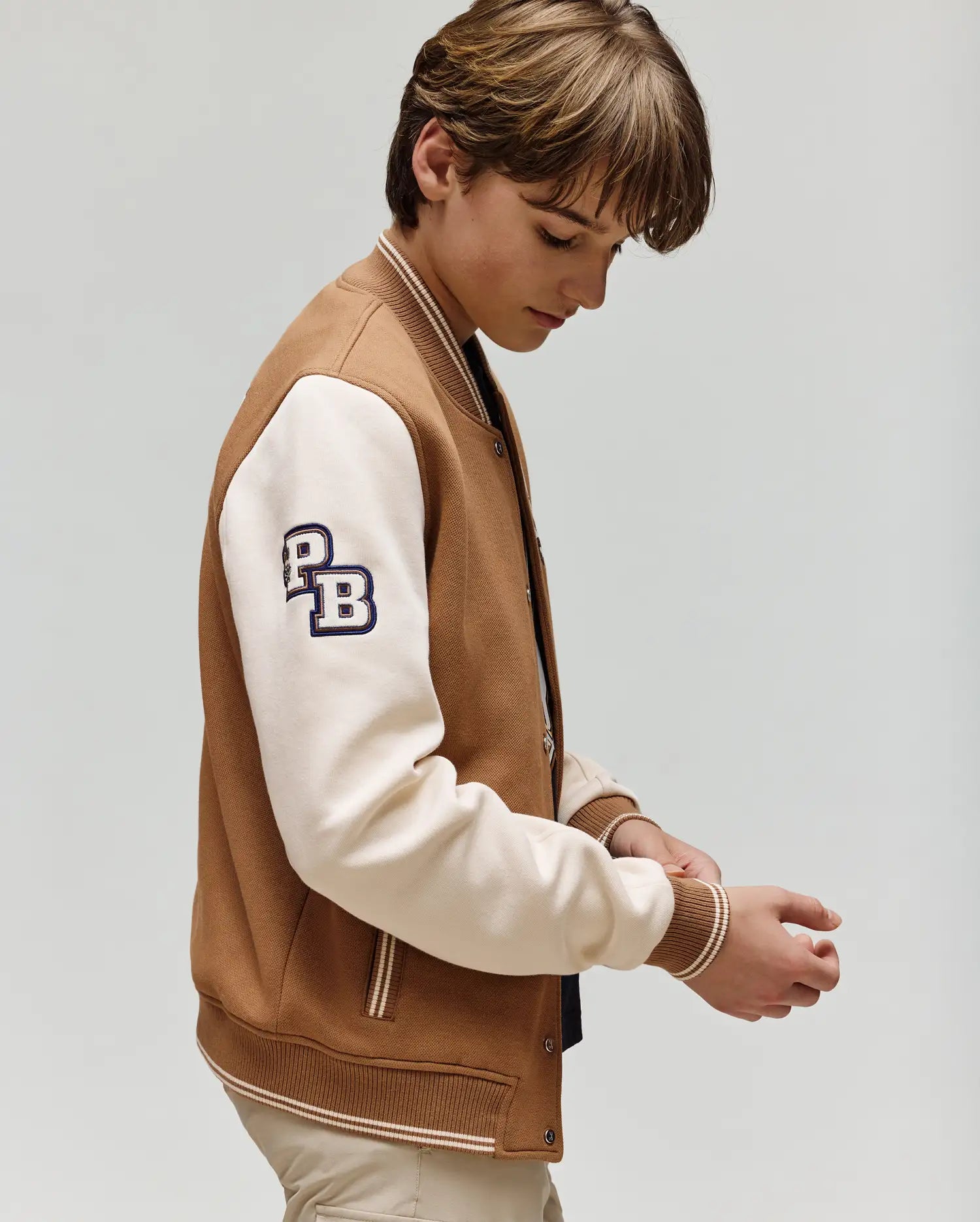 A young person with light brown hair adjusts their cuff while wearing the tan and white Psycho Bunny KIDS WADE KNIT BOMBER (KO1100001) in varsity style, featuring “PB” on the sleeve, against a plain light background.