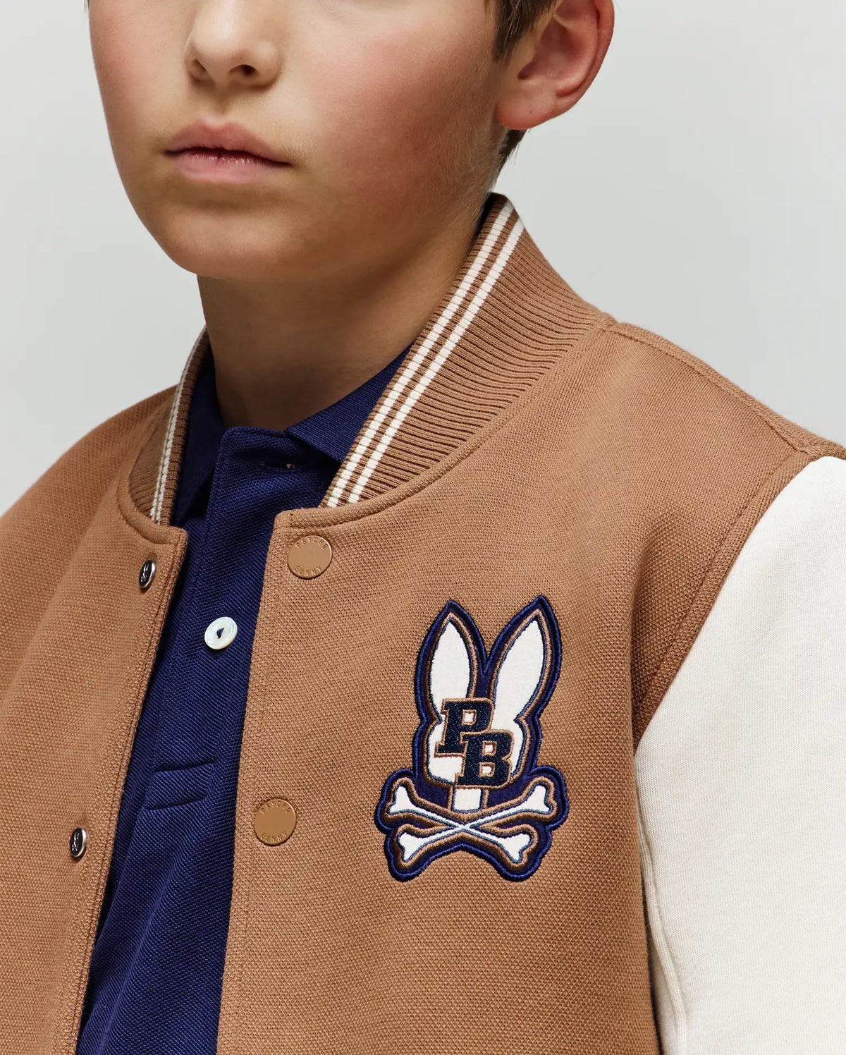 A young boy wears the Psycho Bunny KIDS WADE KNIT BOMBER (KO1100001), a varsity-style cotton blend jacket with a brown and white bunny ears and crossbones logo, layered over a navy collared shirt. Only his lower face is visible.