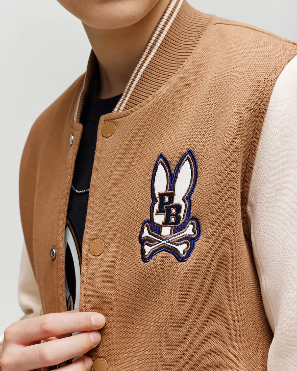 A child wears the Psycho Bunny KIDS WADE KNIT BOMBER (KO1100001), a tan and cream varsity jacket with a bunny head, crossed bones, and "PB" patch on the chest. Made from a premium cotton blend. Only the lower half of their face is visible.