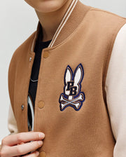 A child wears the Psycho Bunny KIDS WADE KNIT BOMBER (KO1100001), a tan and cream varsity jacket with a bunny head, crossed bones, and "PB" patch on the chest. Made from a premium cotton blend. Only the lower half of their face is visible.