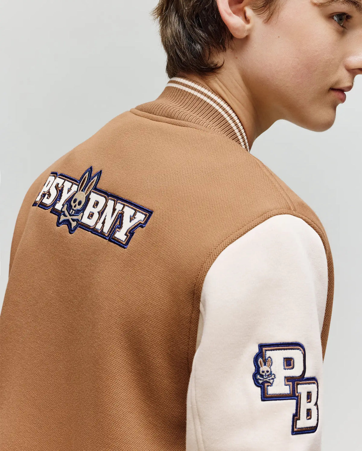 A person is shown from the side, slightly turned away, wearing Psycho Bunny's KIDS WADE KNIT BOMBER (KO1100001)—a brown and cream cotton blend varsity jacket with "PSYBNY" and a rabbit logo on the back and "PB" plus rabbit on the sleeve.