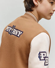 A person is shown from the side, slightly turned away, wearing Psycho Bunny's KIDS WADE KNIT BOMBER (KO1100001)—a brown and cream cotton blend varsity jacket with "PSYBNY" and a rabbit logo on the back and "PB" plus rabbit on the sleeve.