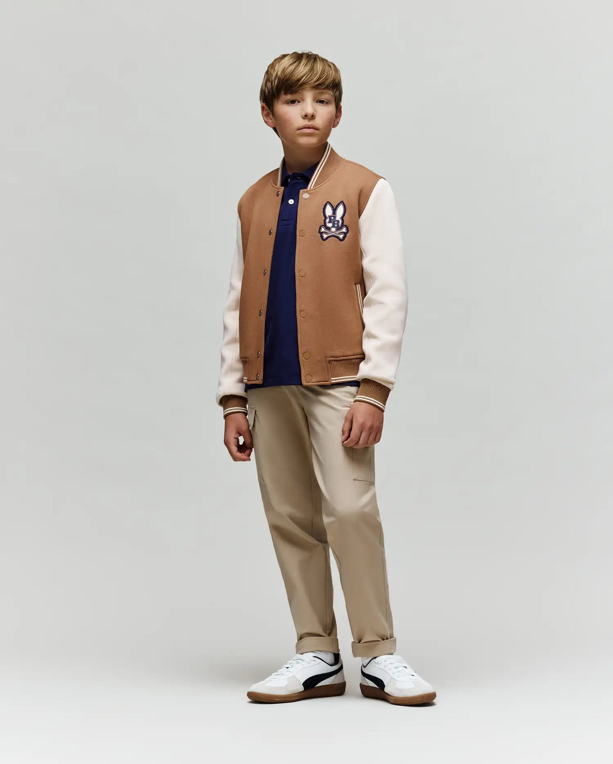 A boy stands slightly to the side, wearing the Psycho Bunny KIDS WADE KNIT BOMBER (KO1100001) in a premium cotton blend, navy polo, beige pants, and white sneakers against a plain light gray background.