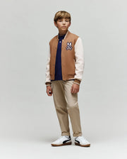 A boy stands slightly to the side, wearing the Psycho Bunny KIDS WADE KNIT BOMBER (KO1100001) in a premium cotton blend, navy polo, beige pants, and white sneakers against a plain light gray background.