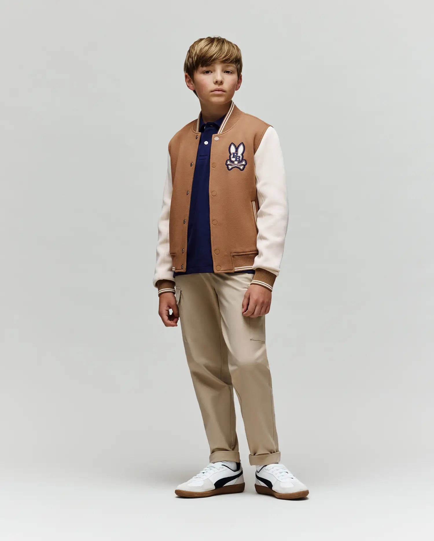 A boy stands slightly to the side, wearing the Psycho Bunny KIDS WADE KNIT BOMBER (KO1100001) in a premium cotton blend, navy polo, beige pants, and white sneakers against a plain light gray background.