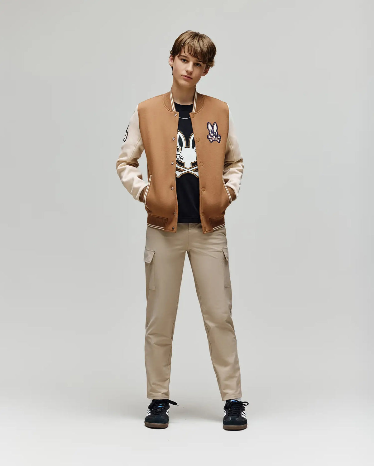 A teen stands against a plain background, hands in pockets, wearing the Psycho Bunny KIDS WADE KNIT BOMBER (KO1100001), beige cargo pants, black sneakers, and a black cartoon graphic T-shirt.