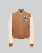 The Psycho Bunny KIDS WADE KNIT BOMBER (KO1100001) is a brown and cream varsity jacket with cream sleeves, brown cuffs and waistband, snap buttons, a bunny patch on the left chest, and a "B" patch on the right sleeve. Made from premium cotton blend. Light gray background.