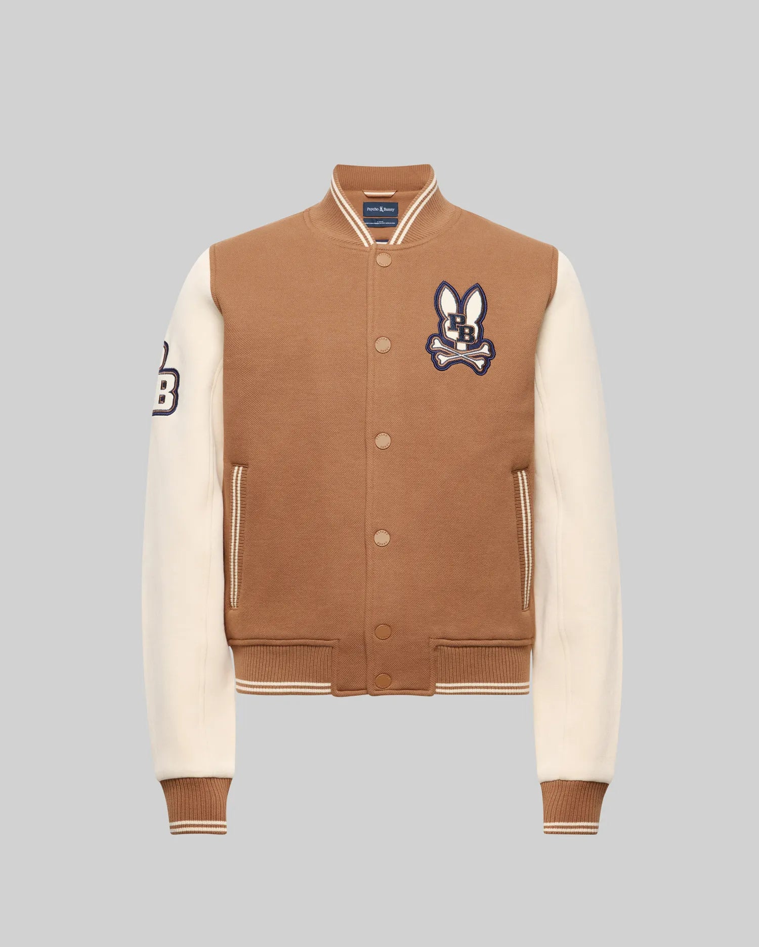 The Psycho Bunny KIDS WADE KNIT BOMBER (KO1100001) is a brown and cream varsity jacket with cream sleeves, brown cuffs and waistband, snap buttons, a bunny patch on the left chest, and a "B" patch on the right sleeve. Made from premium cotton blend. Light gray background.