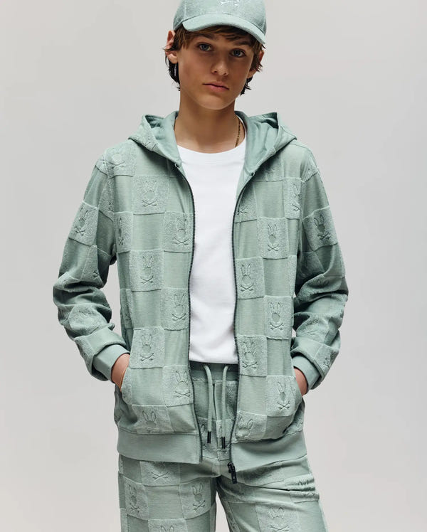 A person with short brown hair and a white t-shirt stands hands-in-pockets, wearing the Psycho Bunny KIDS KELVIN FULL ZIP TOWEL TERRY HOODIE (KT0500004) in light green, matching jogger pants, and a cap. The background is plain gray.
