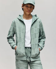 A person with short brown hair and a white t-shirt stands hands-in-pockets, wearing the Psycho Bunny KIDS KELVIN FULL ZIP TOWEL TERRY HOODIE (KT0500004) in light green, matching jogger pants, and a cap. The background is plain gray.