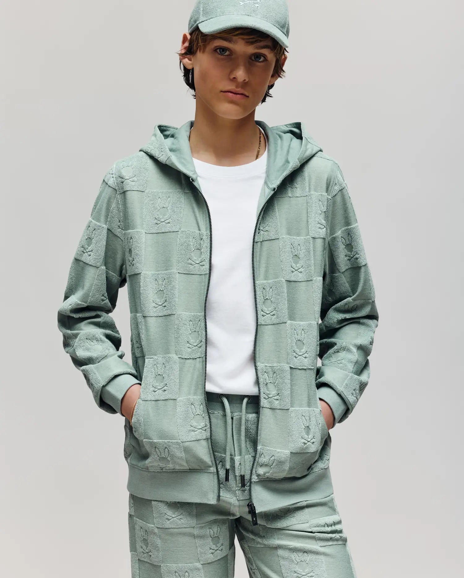 A person with short brown hair and a white t-shirt stands hands-in-pockets, wearing the Psycho Bunny KIDS KELVIN FULL ZIP TOWEL TERRY HOODIE (KT0500004) in light green, matching jogger pants, and a cap. The background is plain gray.