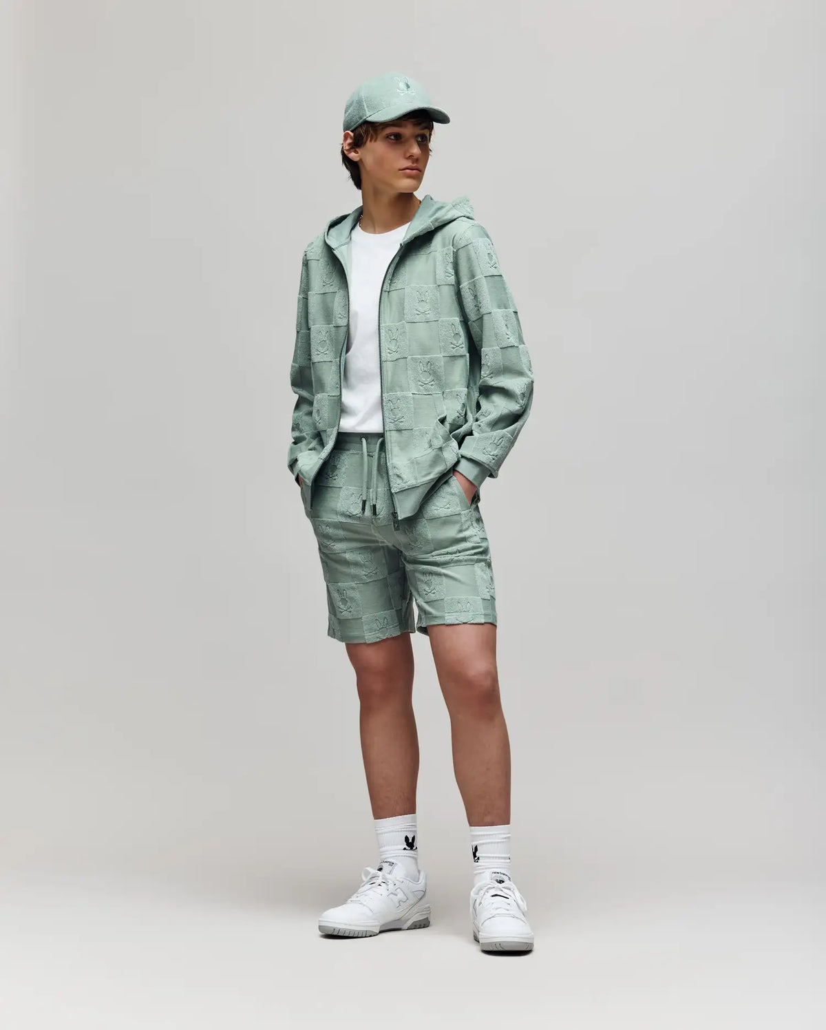A person stands against a plain background wearing the Psycho Bunny KIDS KELVIN FULL ZIP TOWEL TERRY HOODIE (KT0500004) in mint green with matching shorts, a white t-shirt, white socks, and white sneakers. Hands are in the hoodie pockets.