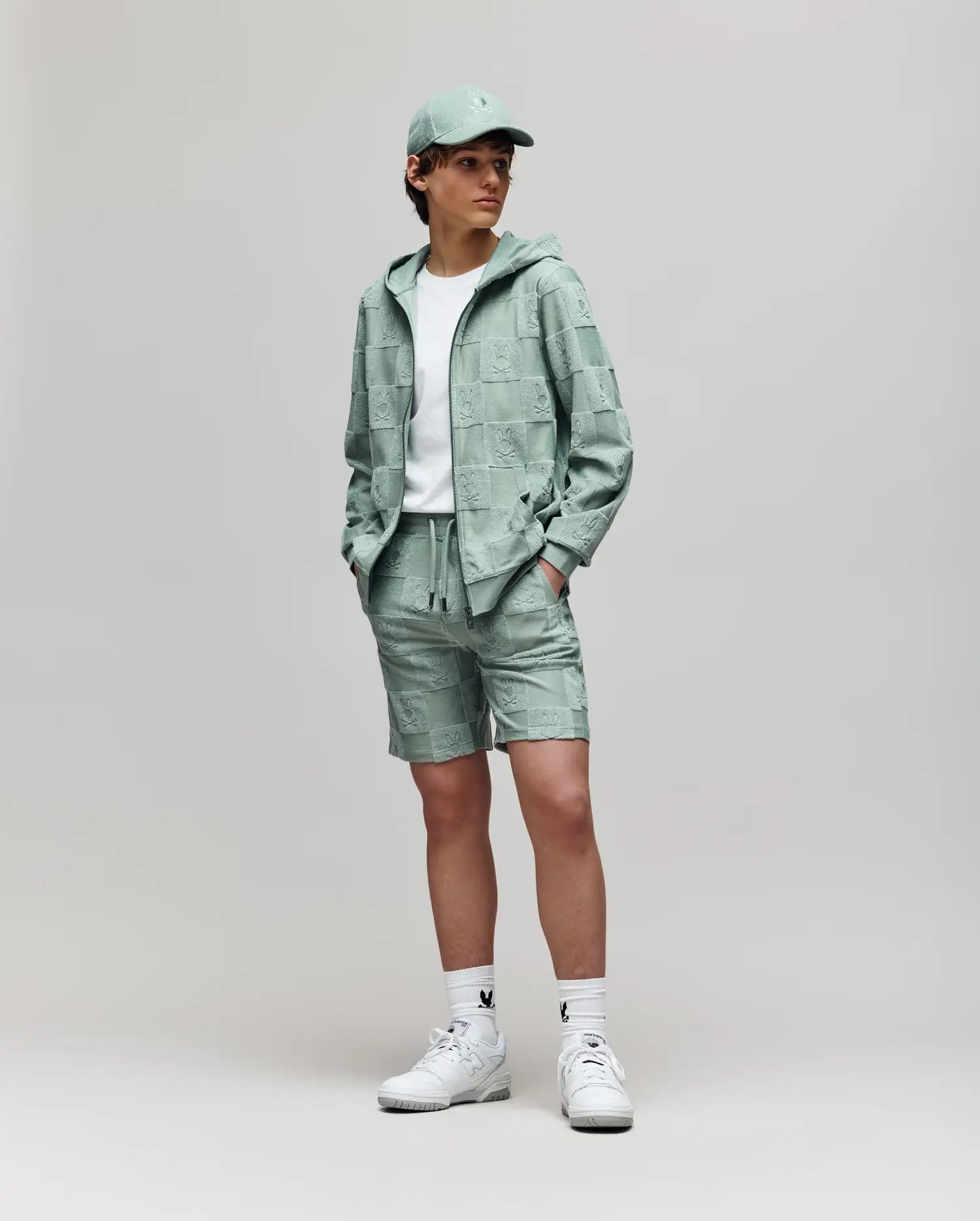 A person stands against a plain background wearing the Psycho Bunny KIDS KELVIN FULL ZIP TOWEL TERRY HOODIE (KT0500004) in mint green with matching shorts, a white t-shirt, white socks, and white sneakers. Hands are in the hoodie pockets.