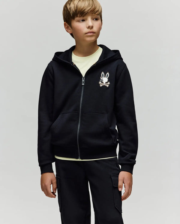 A young boy wears the Psycho Bunny KIDS WADE CORD PATCH ZIP HOODIE (KT0500016) with an embroidered Bunny skull and crossbones on the chest, a pale tee, and black cargo pants against a plain light gray background.