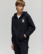 A young person with light brown hair wears the Psycho Bunny KIDS WADE CORD PATCH ZIP HOODIE (KT0500016), featuring an embroidered Bunny and crossbones logo, over a white T-shirt and black pants, standing against a light gray background.