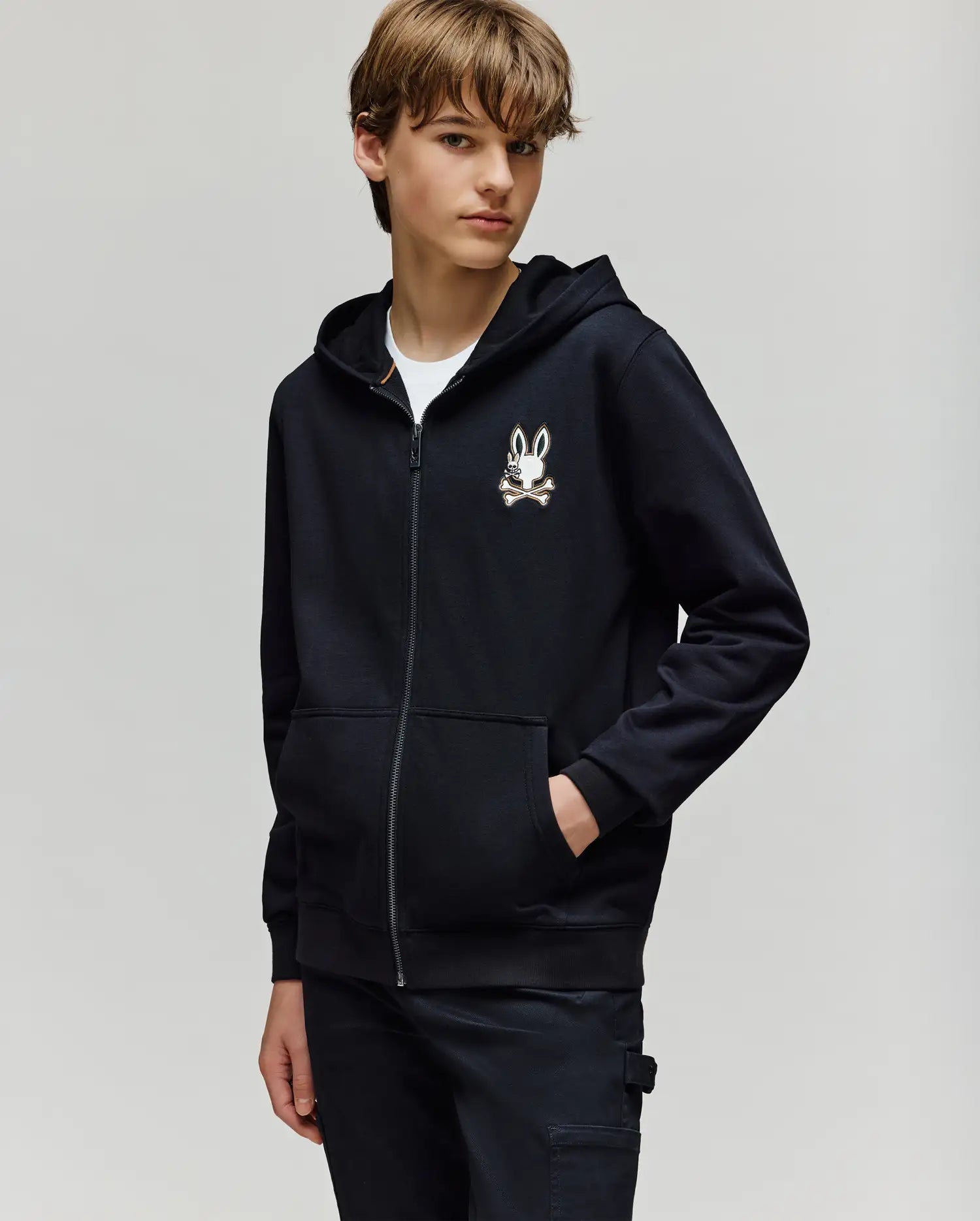 A young person with light brown hair wears the Psycho Bunny KIDS WADE CORD PATCH ZIP HOODIE (KT0500016), featuring an embroidered Bunny and crossbones logo, over a white T-shirt and black pants, standing against a light gray background.