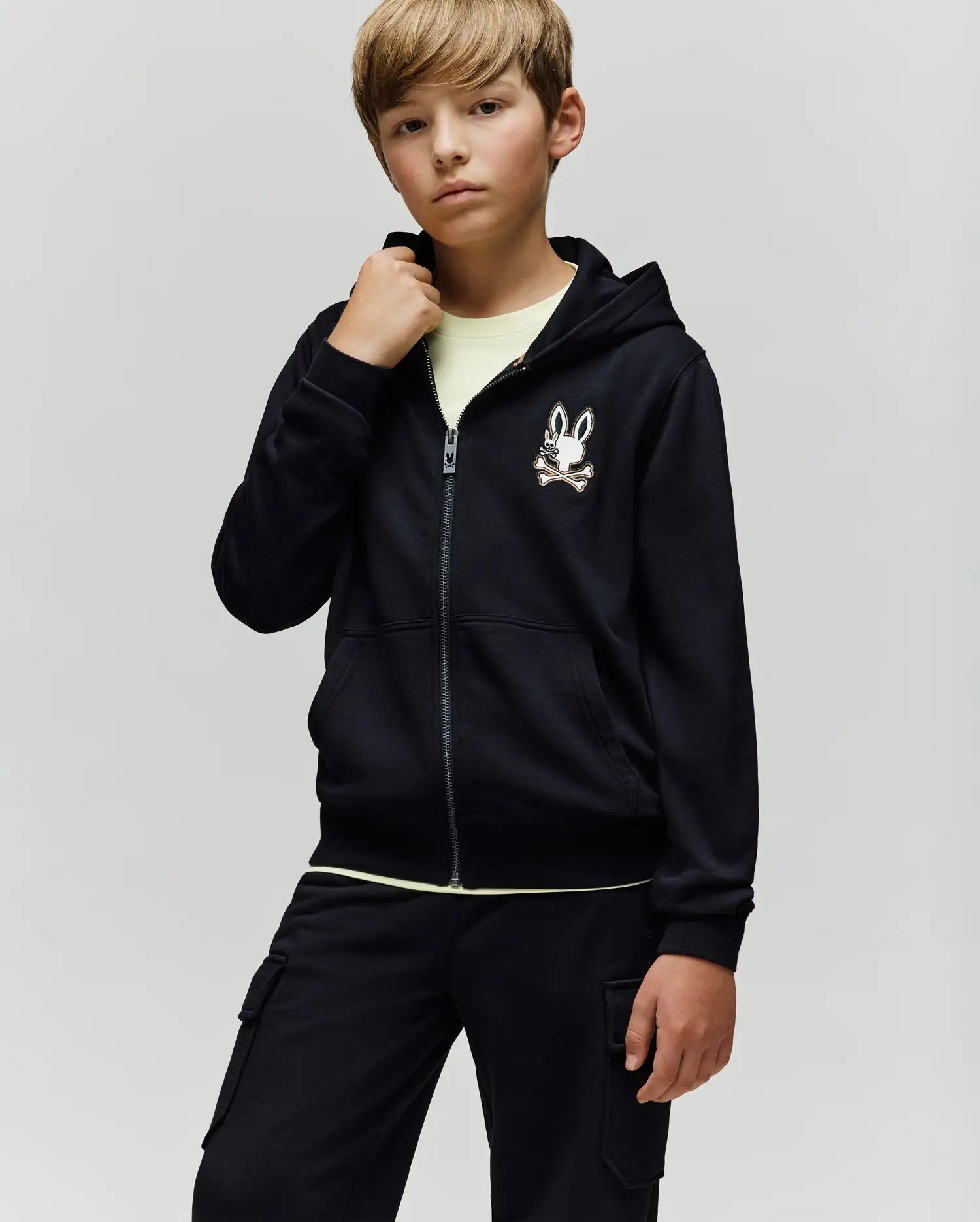 A young boy with light brown hair wears the Psycho Bunny KIDS WADE CORD PATCH ZIP HOODIE (KT0500016) in black, paired with a light shirt and black cargo pants, standing against a plain light background.