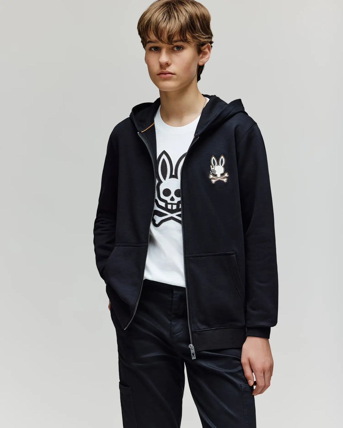 A young person with short, light brown hair wears the Psycho Bunny KIDS WADE CORD PATCH ZIP HOODIE (KT0500016) over a white tee with an embroidered Bunny and crossbones, one hand in their pocket, standing against a plain light background.