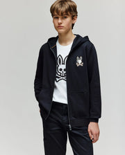 A young person with short, light brown hair wears the Psycho Bunny KIDS WADE CORD PATCH ZIP HOODIE (KT0500016) over a white tee with an embroidered Bunny and crossbones, one hand in their pocket, standing against a plain light background.