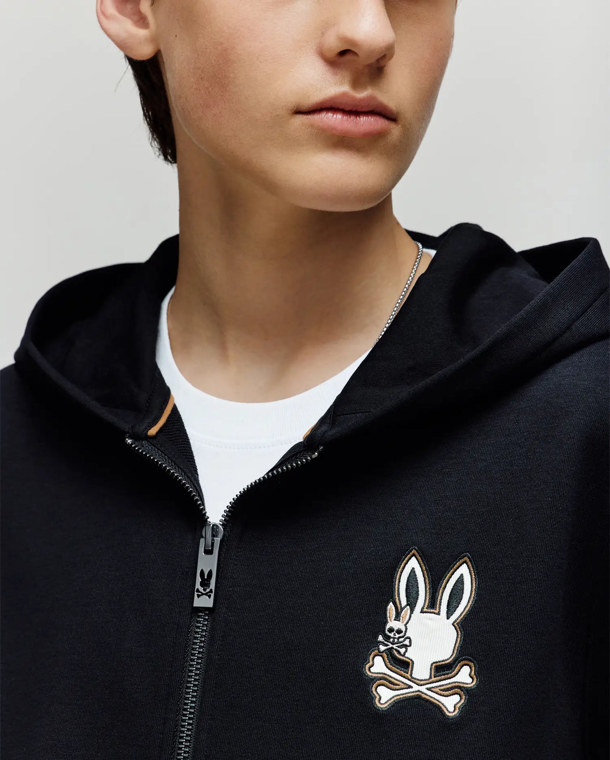 A child wears the Psycho Bunny KIDS WADE CORD PATCH ZIP HOODIE (KT0500016), a black premium cotton zip-hoodie with an embroidered Bunny logo and crossbones on the chest, layered over a white t-shirt. Their face is partially visible.