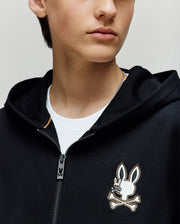 A child wears the Psycho Bunny KIDS WADE CORD PATCH ZIP HOODIE (KT0500016), a black premium cotton zip-hoodie with an embroidered Bunny logo and crossbones on the chest, layered over a white t-shirt. Their face is partially visible.