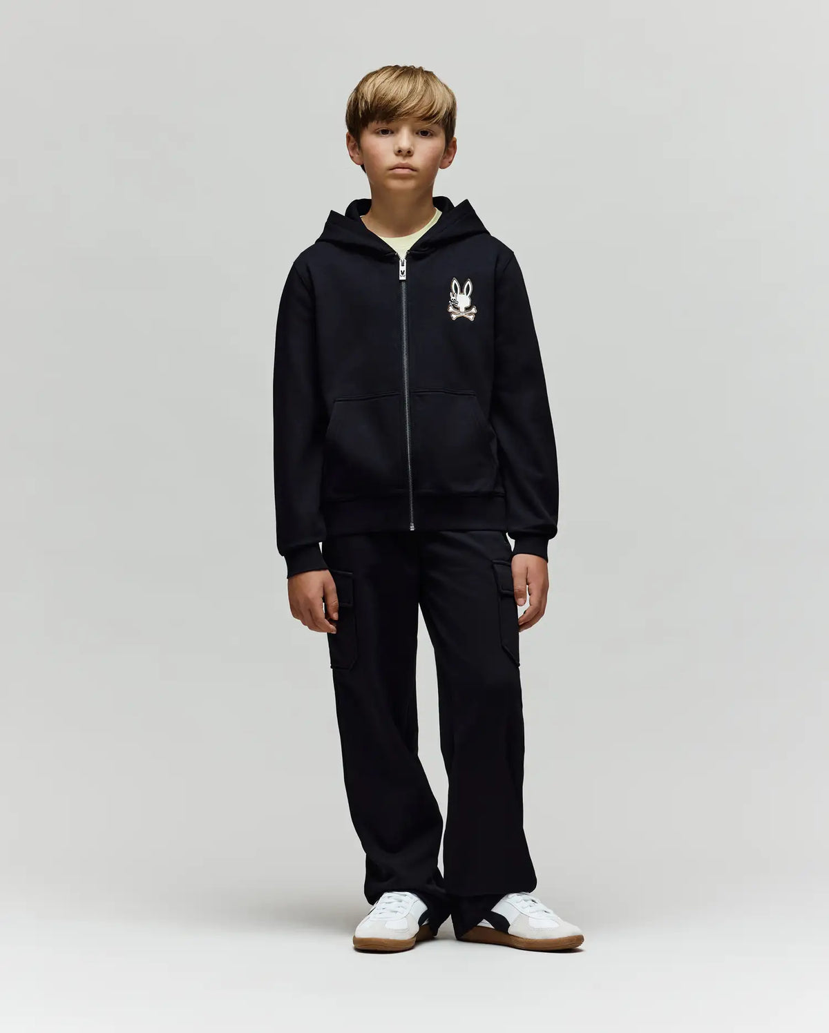 A blonde boy stands facing forward in a Psycho Bunny KIDS WADE CORD PATCH ZIP HOODIE (KT0500016) with an embroidered Bunny graphic on the left chest, black cargo pants, and white sneakers against a plain light gray background.