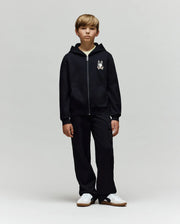 A blonde boy stands facing forward in a Psycho Bunny KIDS WADE CORD PATCH ZIP HOODIE (KT0500016) with an embroidered Bunny graphic on the left chest, black cargo pants, and white sneakers against a plain light gray background.