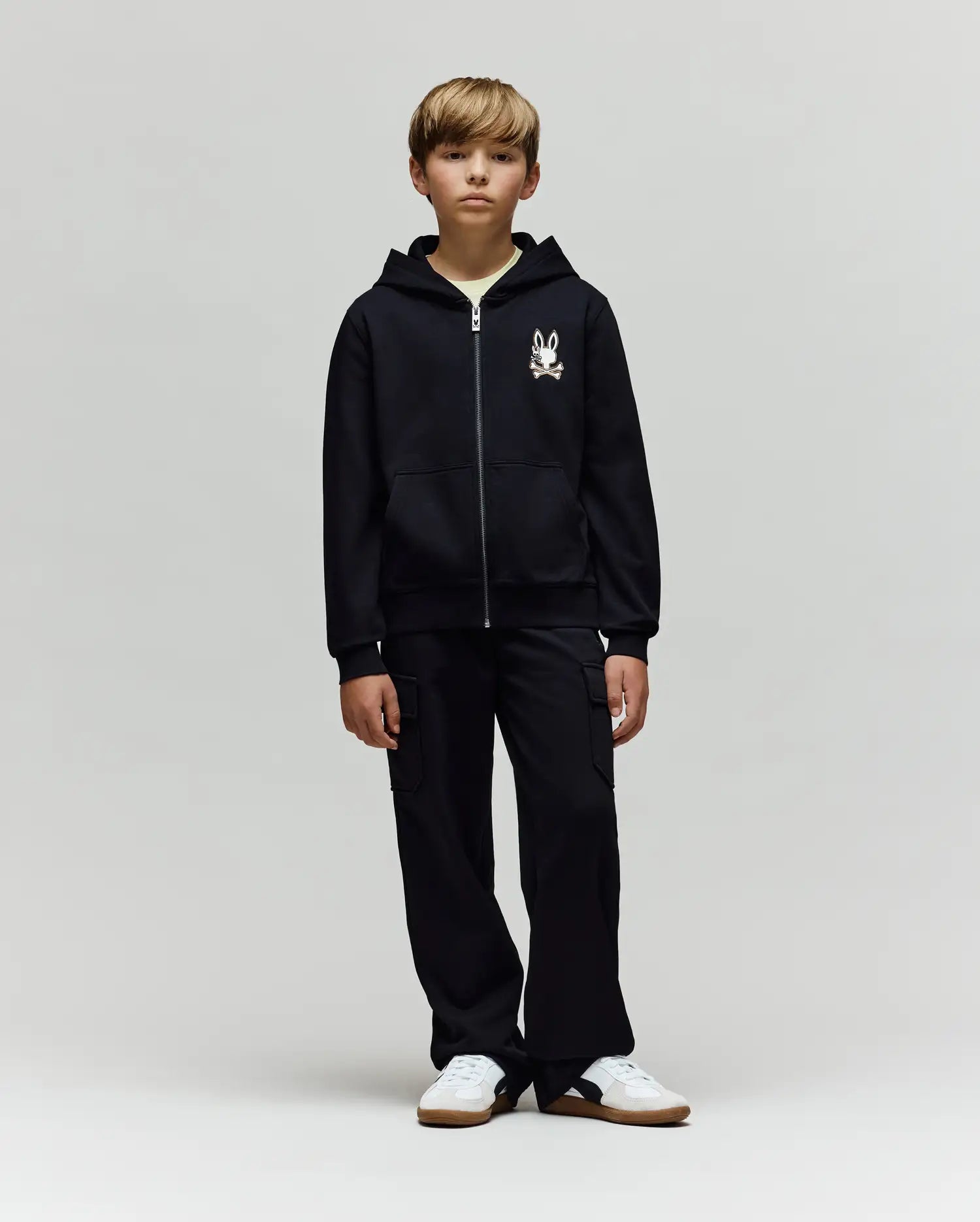 A blonde boy stands facing forward in a Psycho Bunny KIDS WADE CORD PATCH ZIP HOODIE (KT0500016) with an embroidered Bunny graphic on the left chest, black cargo pants, and white sneakers against a plain light gray background.