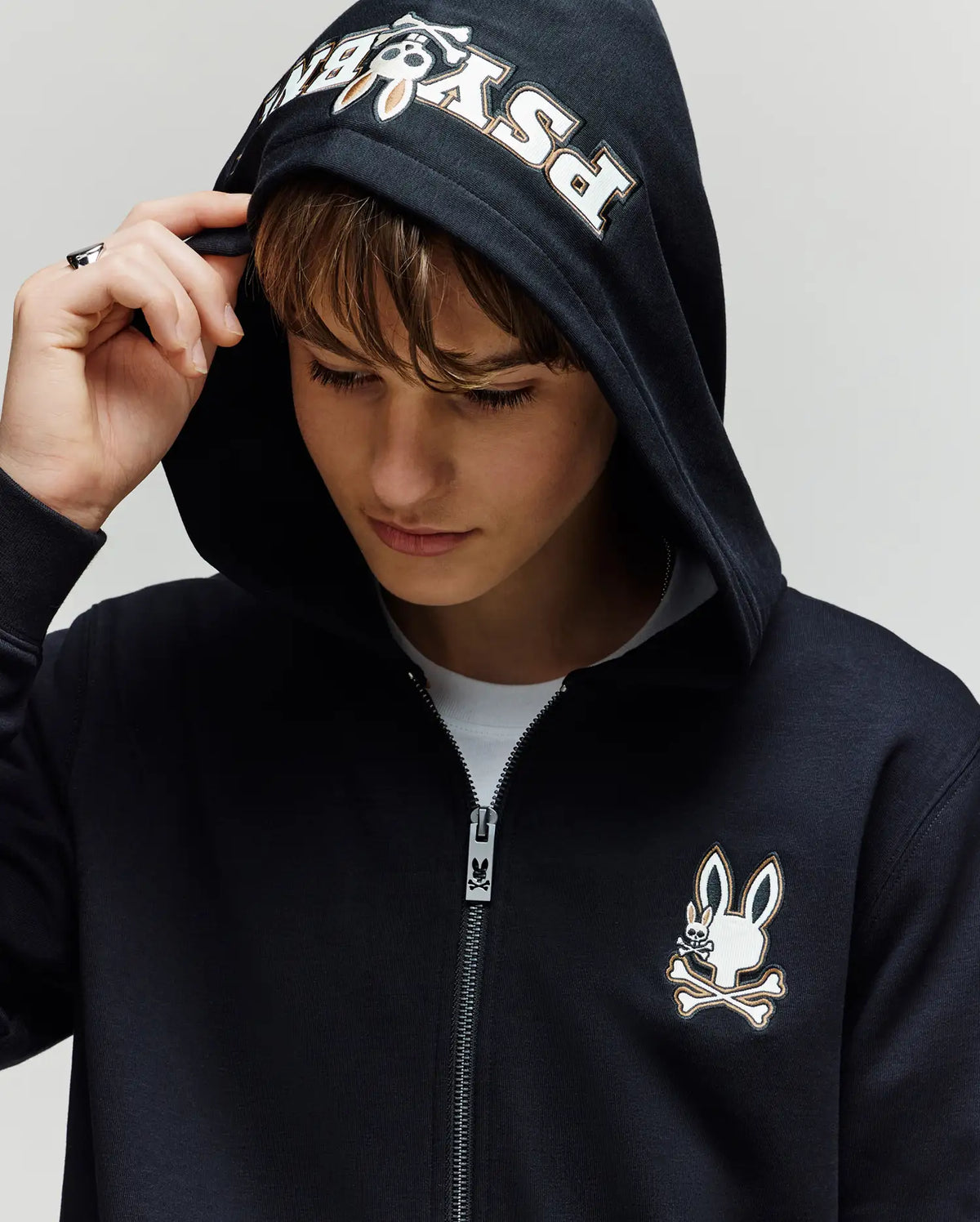 A young person in the Psycho Bunny KIDS WADE CORD PATCH ZIP HOODIE (KT0500016), featuring "Psycho" on the hood and an embroidered Bunny skull-and-crossbones logo on the chest, looks down holding the hood’s edge.