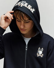 A young person in the Psycho Bunny KIDS WADE CORD PATCH ZIP HOODIE (KT0500016), featuring "Psycho" on the hood and an embroidered Bunny skull-and-crossbones logo on the chest, looks down holding the hood’s edge.