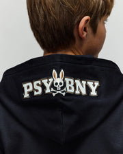 A person seen from behind wears the Psycho Bunny KIDS WADE CORD PATCH ZIP HOODIE (KT0500016) with "PSY BNY" text and an embroidered Bunny skull-and-crossbones logo featuring a rabbit head on the hood.