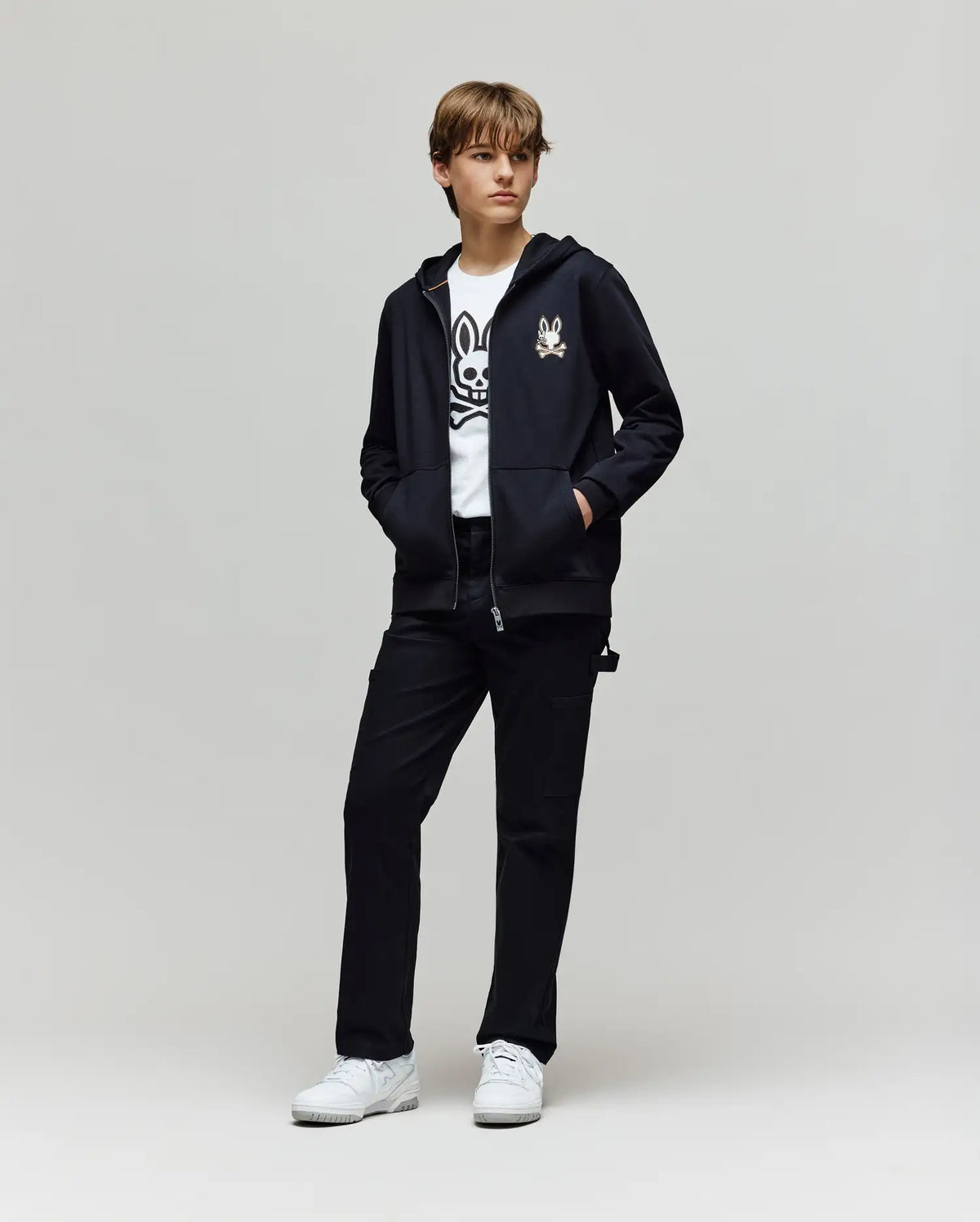 A young person stands against a plain backdrop, hands in pockets, wearing Psycho Bunny’s KIDS WADE CORD PATCH ZIP HOODIE (KT0500016) in black, featuring an embroidered Bunny logo, paired with a graphic white tee, black pants, and white sneakers.