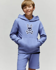 A smiling boy with light brown hair wears the Psycho Bunny KIDS RYDER BUNNY HOODIE (KT0500018), featuring an embroidered Bunny patch, standing hands-in-pockets against a plain background—the perfect loungewear look.