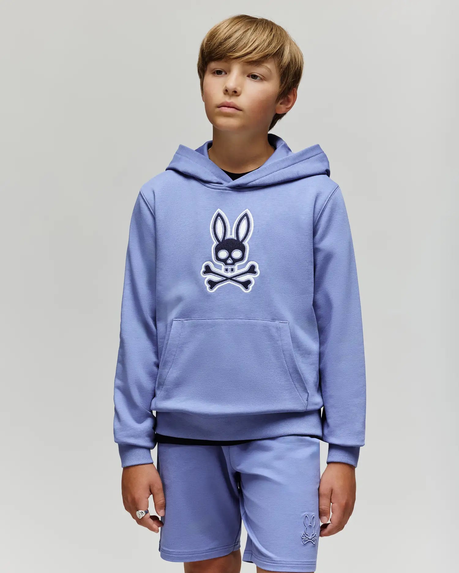 A young boy with light brown hair wears the Psycho Bunny KIDS RYDER BUNNY HOODIE (KT0500018) and matching shorts, standing against a plain light gray background—perfect for relaxed days.
