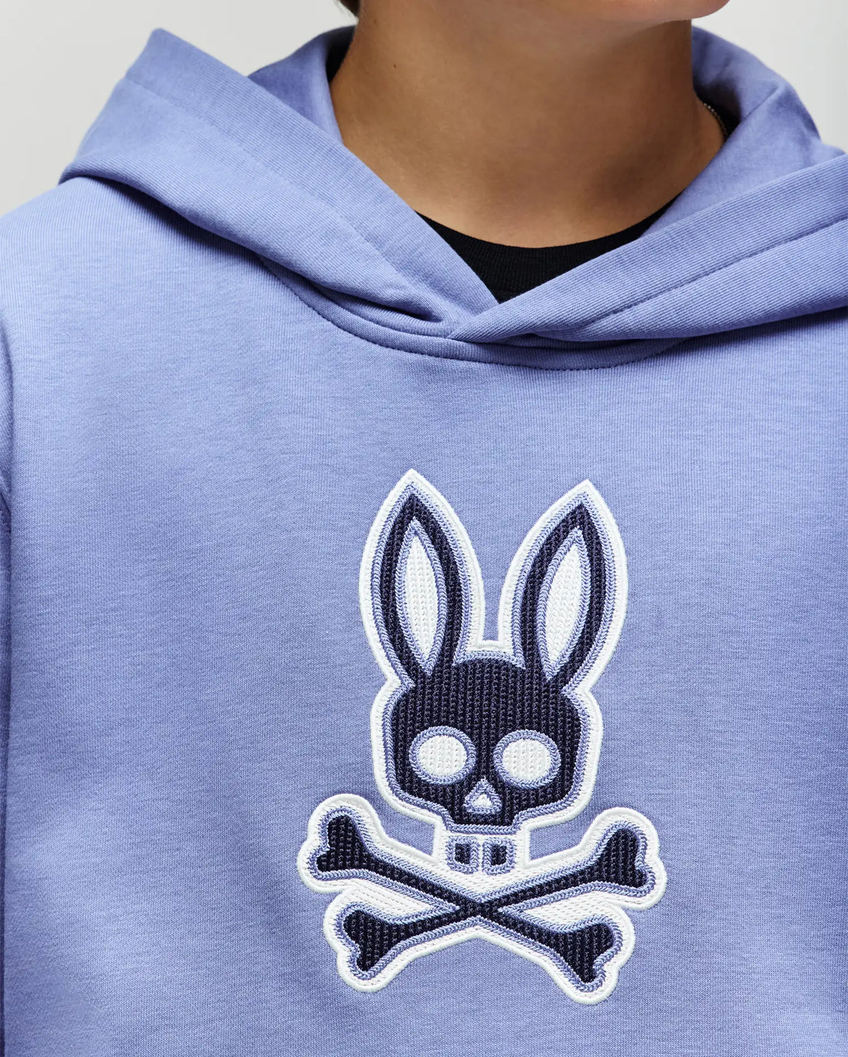 A person wears a Psycho Bunny KIDS RYDER BUNNY HOODIE (KT0500018) in light purple, featuring a large front skull and crossbones graphic with bunny face and ears. Only the lower face and upper torso are visible, making this loungewear unique.