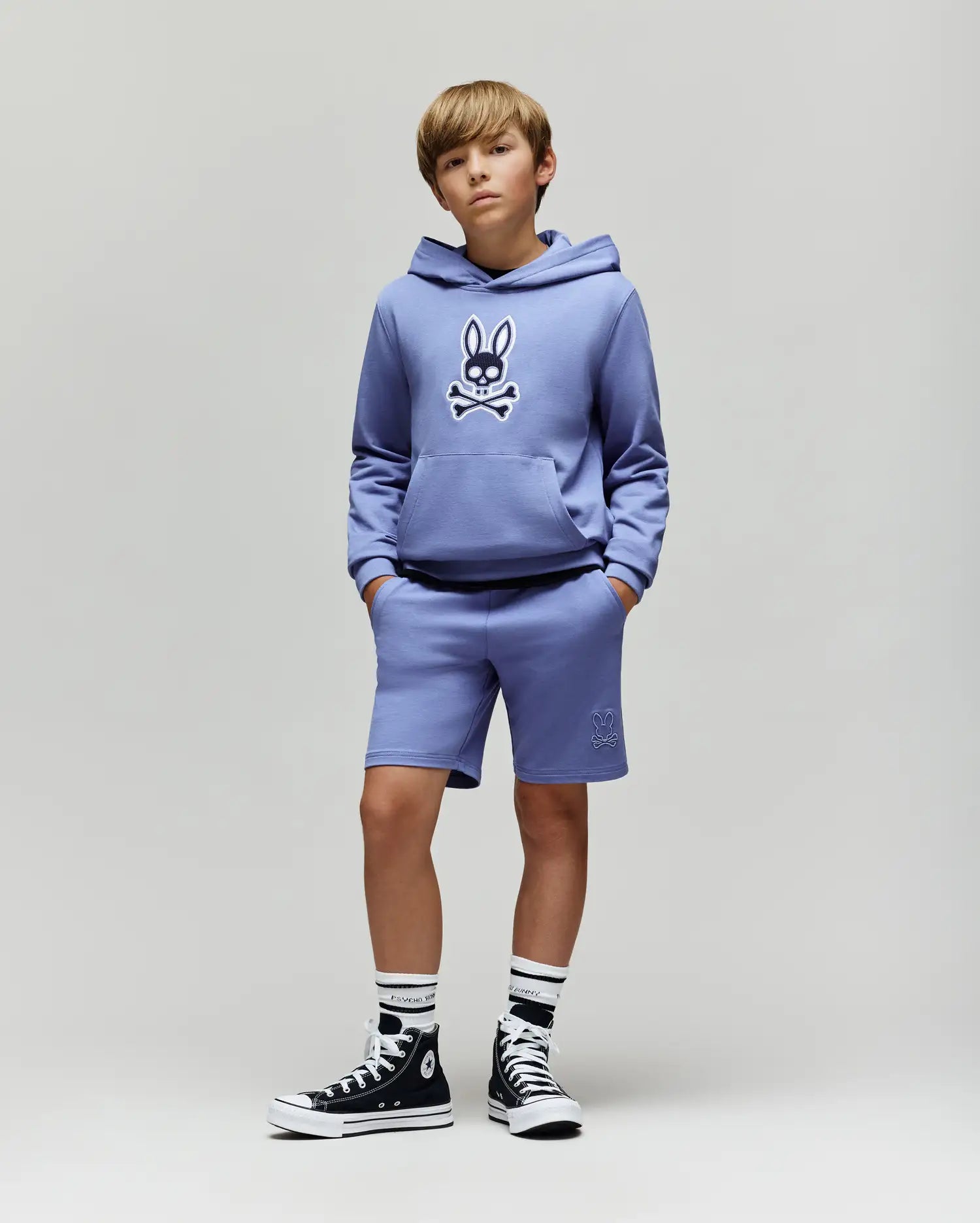 A boy stands against a light background wearing the Psycho Bunny KIDS RYDER BUNNY HOODIE (KT0500018) with an embroidered Bunny patch, matching shorts, striped socks, and black high-top sneakers—a relaxed and stylish look.