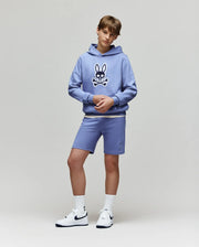 A teen boy stands against a plain background wearing the Psycho Bunny KIDS RYDER BUNNY HOODIE (KT0500018) in lavender with an embroidered Bunny patch, matching shorts, white crew socks, and white Nike sneakers.