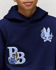 A person wears the Psycho Bunny KIDS AUGUSTE HOODIE (KT0500019), a regular fit dark blue cotton fleece hoodie featuring two embroidered patches: a stylized “PB” with a blue rose and a bunny head with crossbones, blue flowers, and chenille floral details.