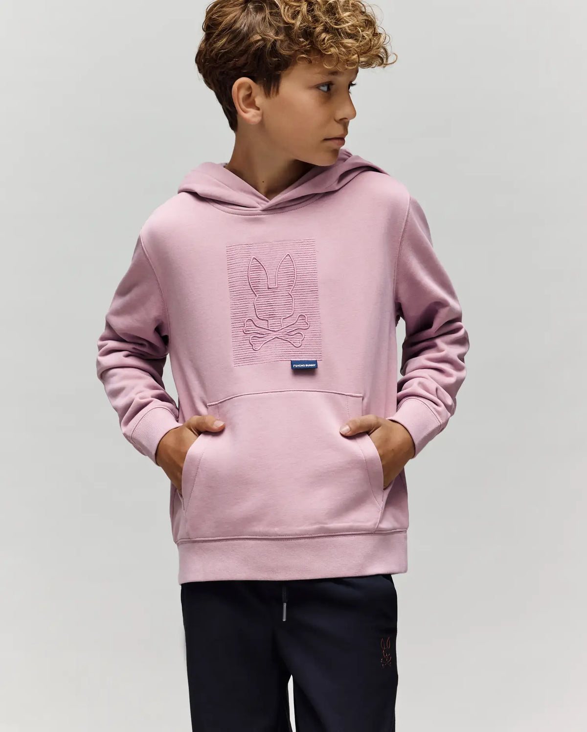 A young boy with curly hair wears the Psycho Bunny KIDS GRANT HOODIE - KT0500020 in light purple, featuring an embroidered bunny and crossbones. He stands with hands in the front pocket, looking right against a plain background.